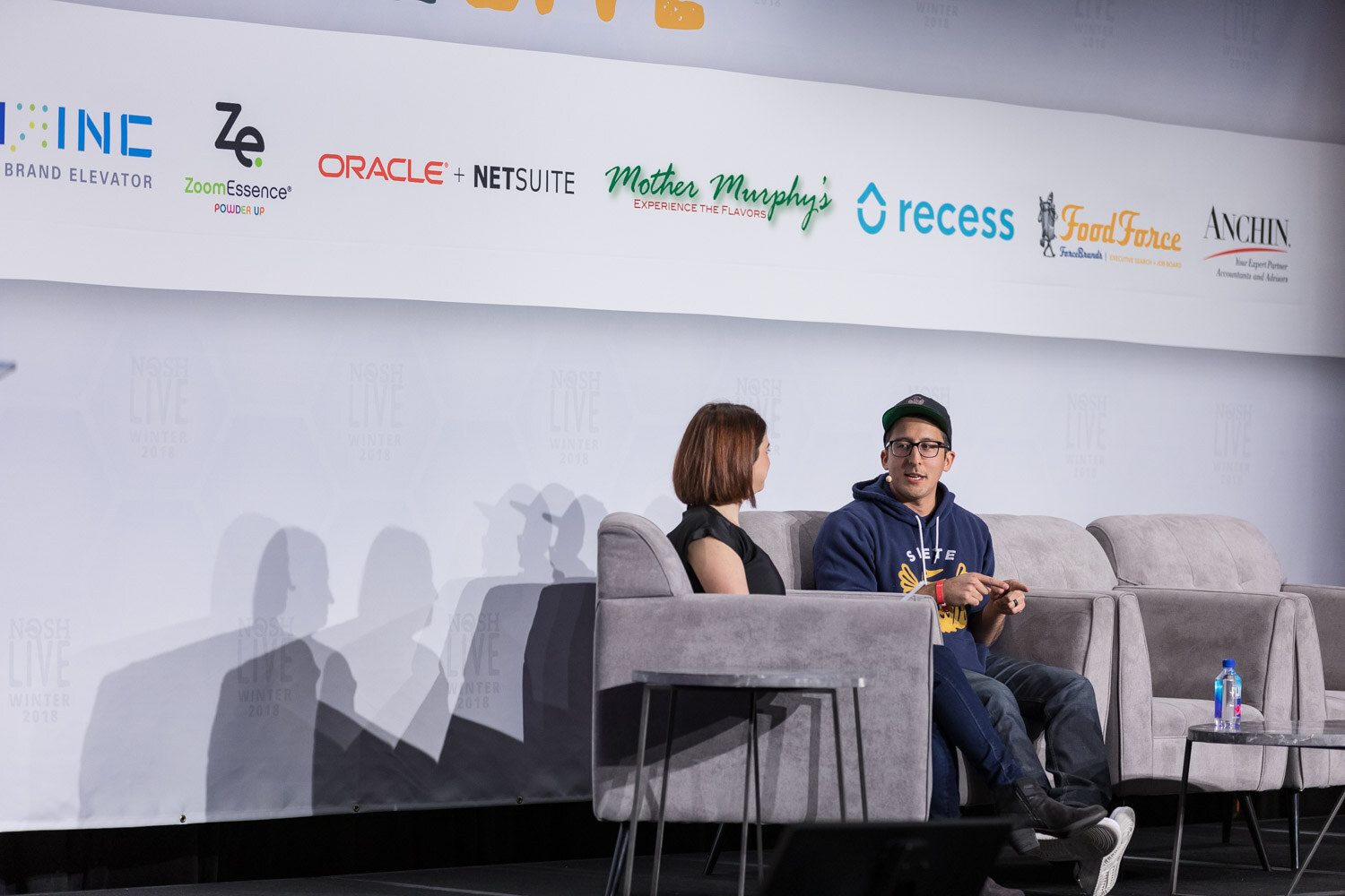 A wide view of two people talking in armchairs on stage under a Nosh Live banner with sponsor logos.