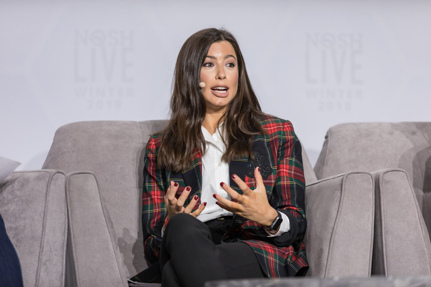 A woman in a red plaid blazer speaks with her hands open on the main stage.
