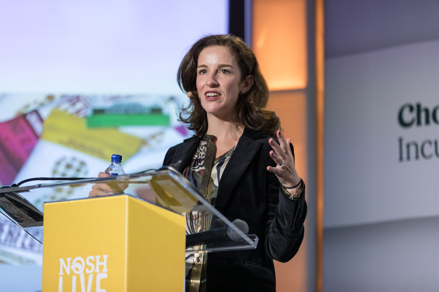 A woman in a black blazer speaks at the yellow Nosh Live lectern with a colorful slide behind her.