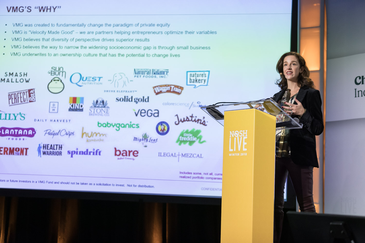 A presenter in a black blazer stands at the lectern in front of a slide titled VMG's Why.