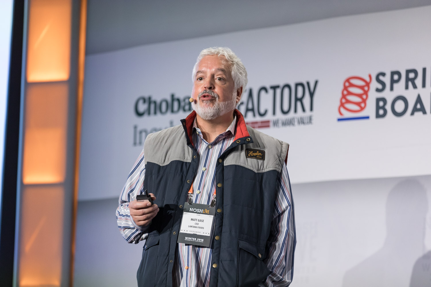 A man in a navy vest and striped shirt presents on the main stage with a clicker.