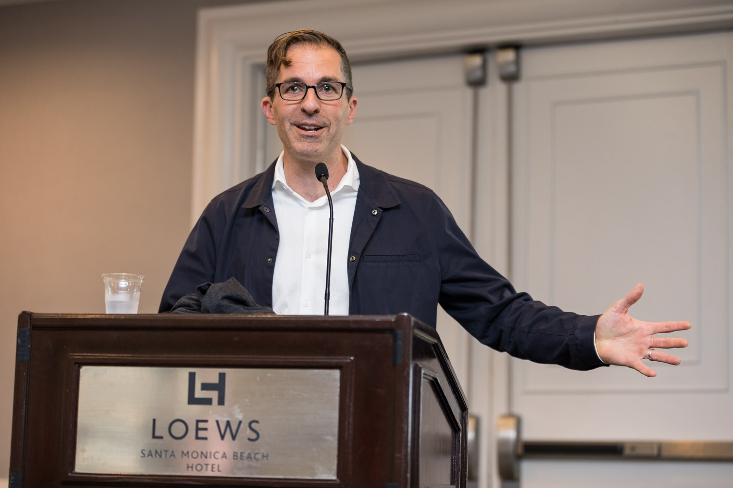 A man in a navy jacket speaks at a lectern marked Loews Santa Monica Beach Hotel during Breakout Session 2.