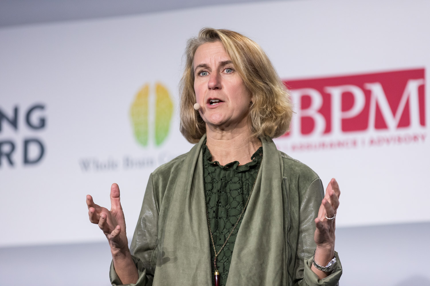 A woman in a green jacket gestures with both hands while speaking on the main stage.