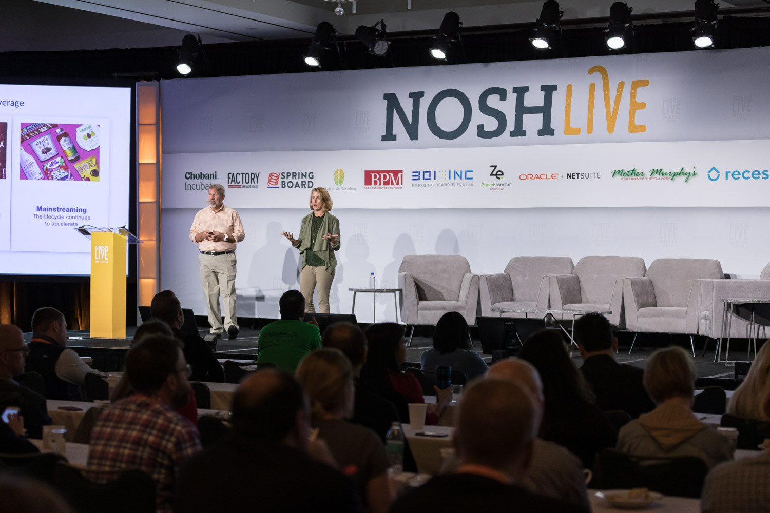 Two presenters stand on the main stage in front of a seated audience.
