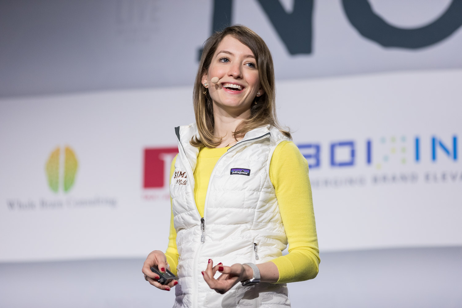 A smiling woman in a white vest and yellow top presents on the main stage.