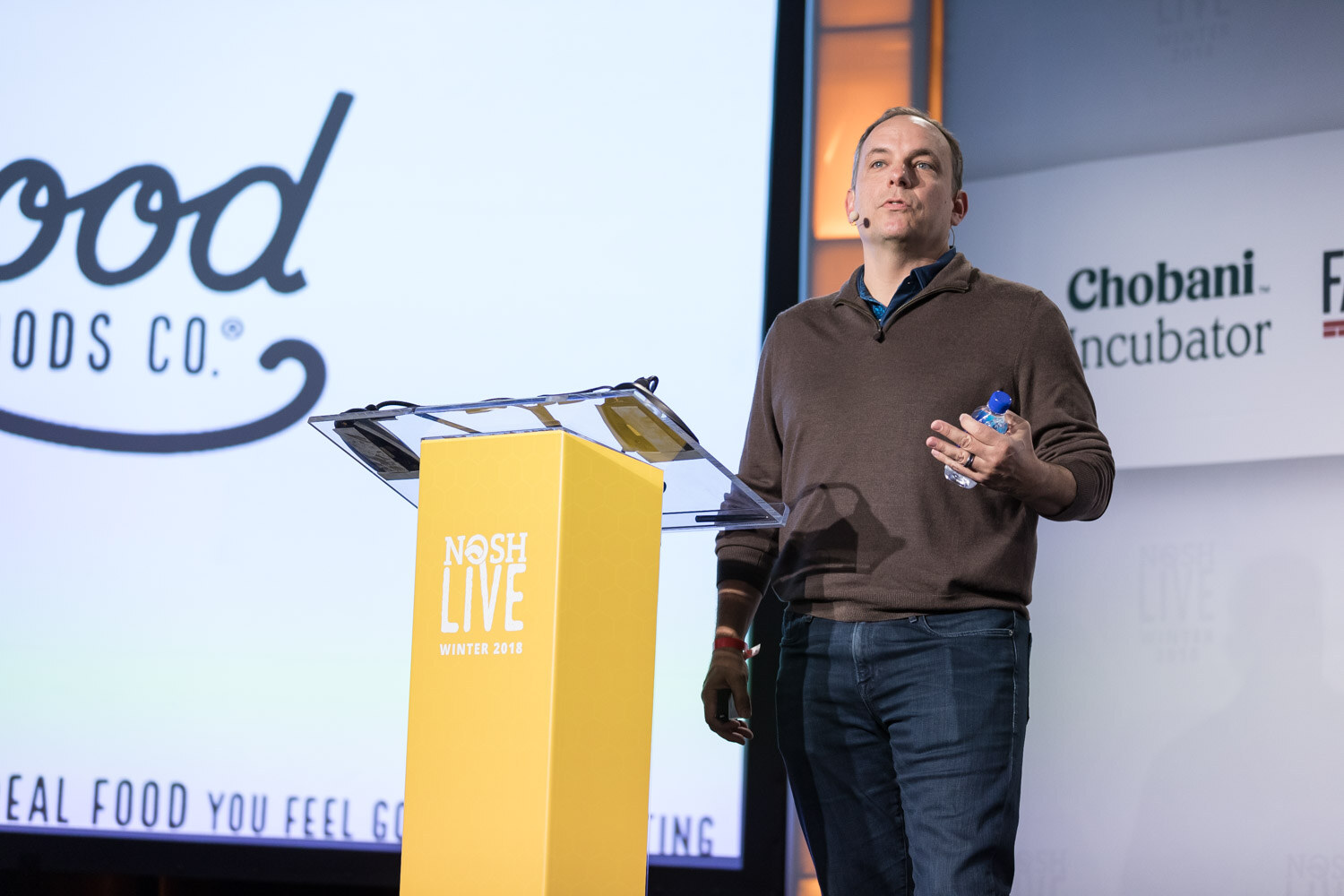 A presenter in a brown sweater holding a water bottle speaks at the lectern beside a slide with a food brand logo.