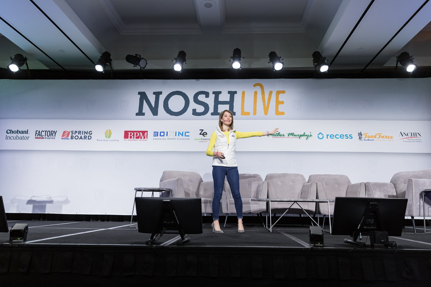 A woman in a white vest and yellow top stands alone on the main stage under the Nosh Live banner.