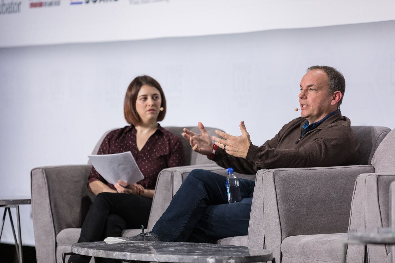 A man in a brown sweater and a woman holding papers talk in armchairs on the main stage.