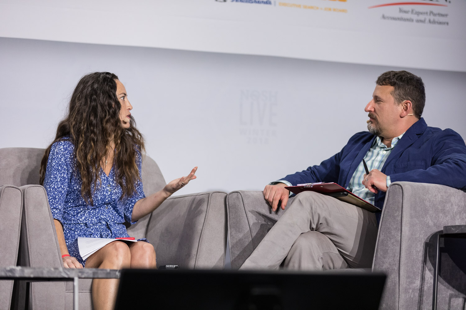 A woman in a blue dress and a man in a blue blazer talk in armchairs on the main stage.