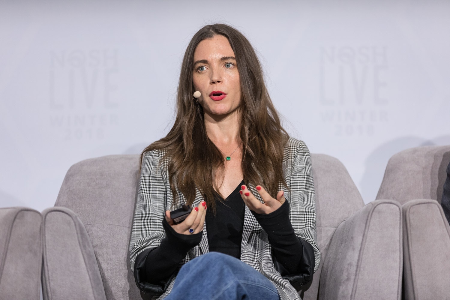 A woman in a gray plaid blazer gestures while speaking in an armchair on the main stage.