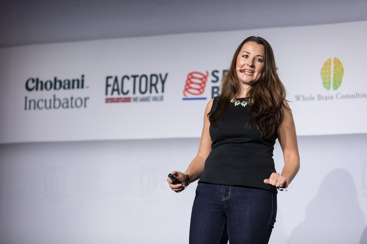 A woman in a black sleeveless top holds a clicker and speaks in front of Chobani Incubator and Factory signs.