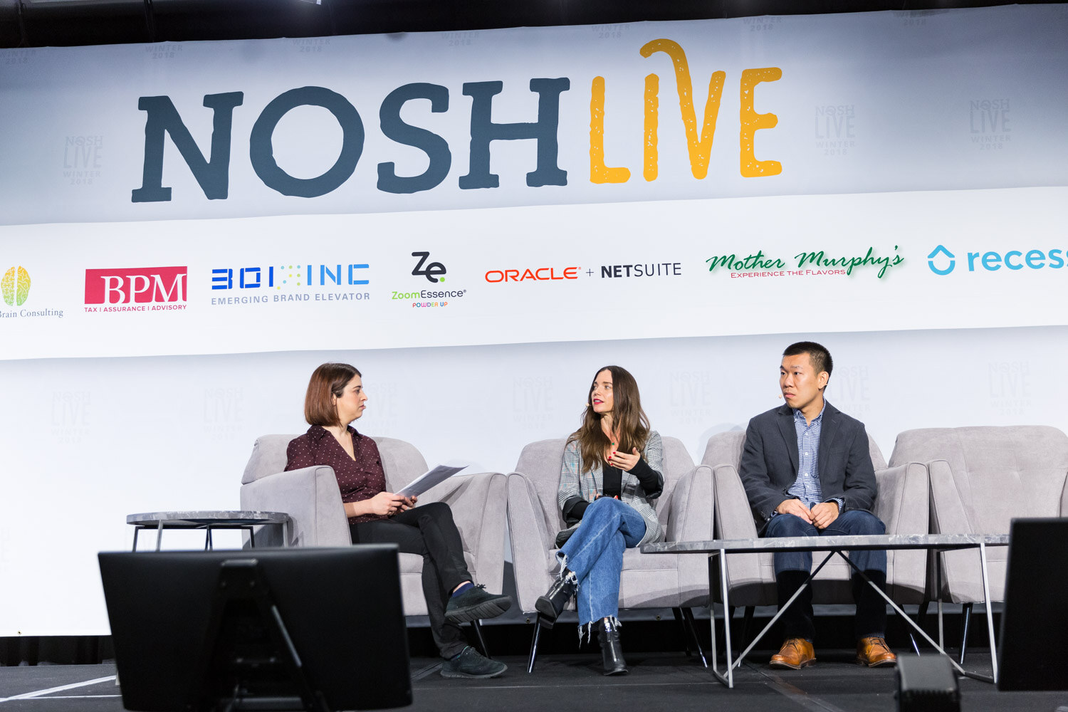Three people sit in gray armchairs on the main stage under the Nosh Live banner.