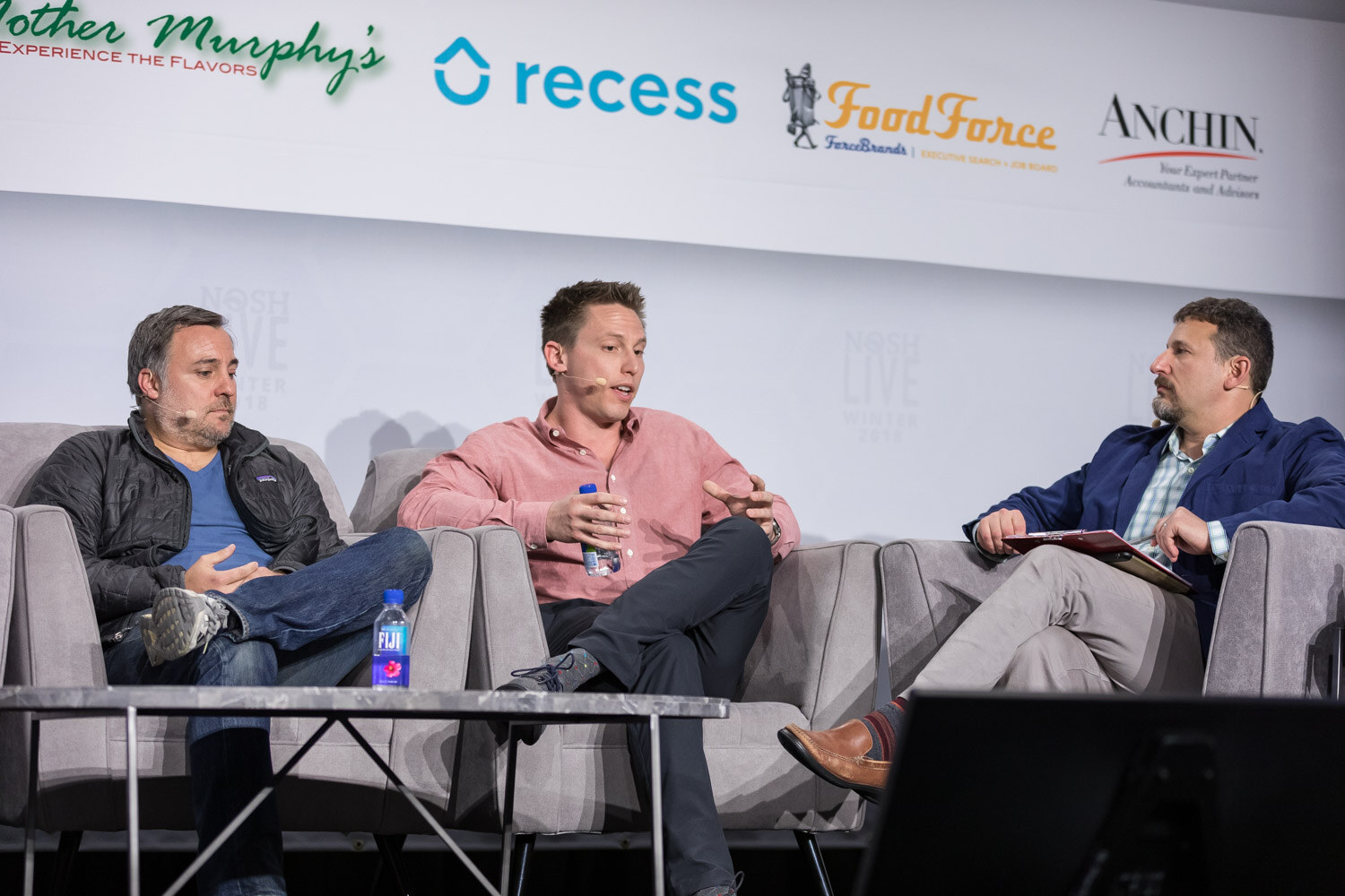 Three panelists sit on gray couches on the main stage under sponsor logos.