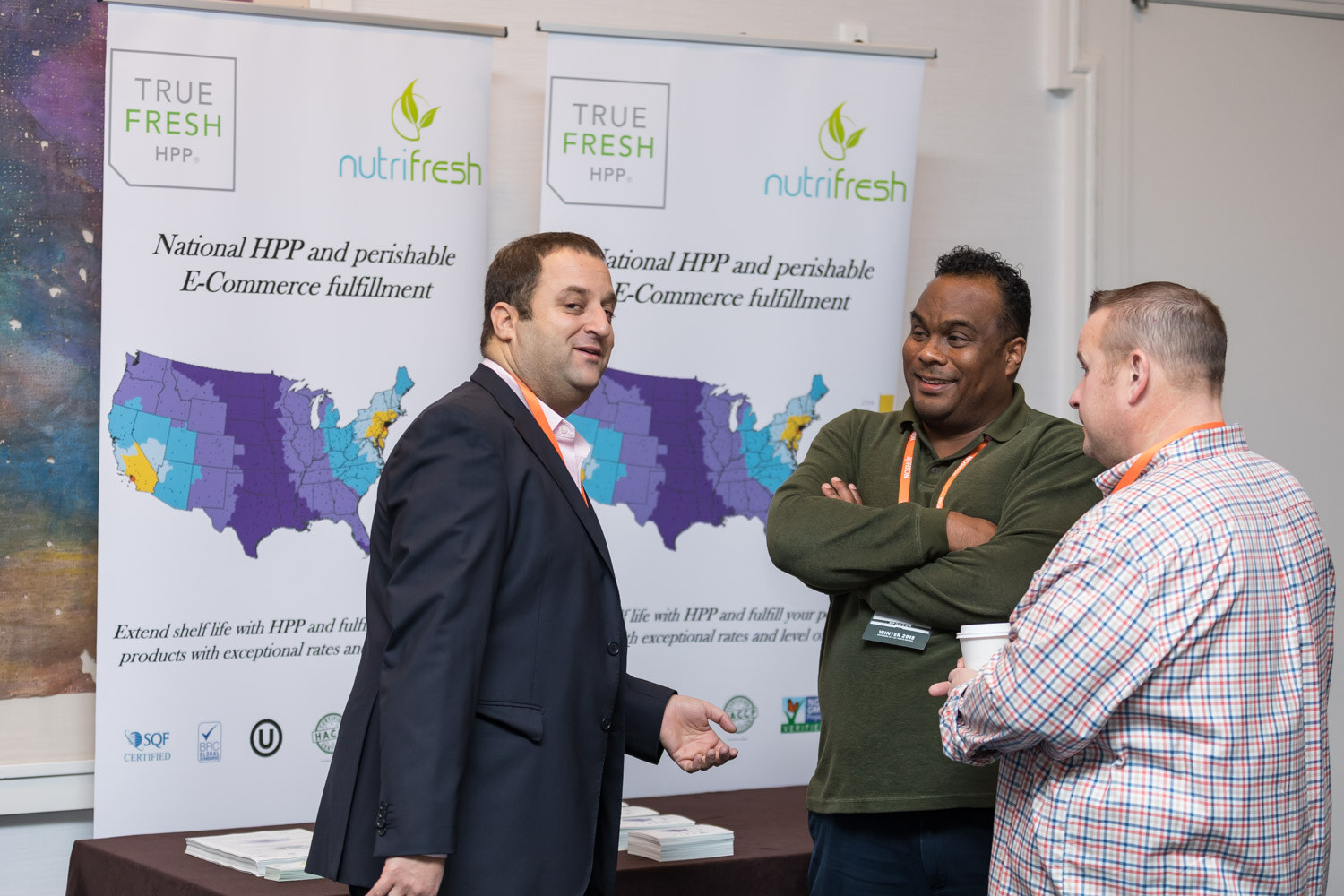 Two men talk in front of True Fresh and Nutrifresh banners at a tabletop exhibit.