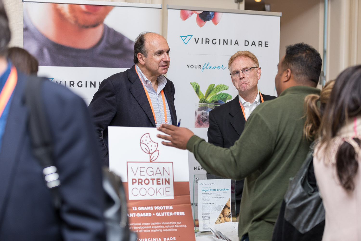 Attendees talk at a Virginia Dare booth with a Vegan Protein Cookie sign.