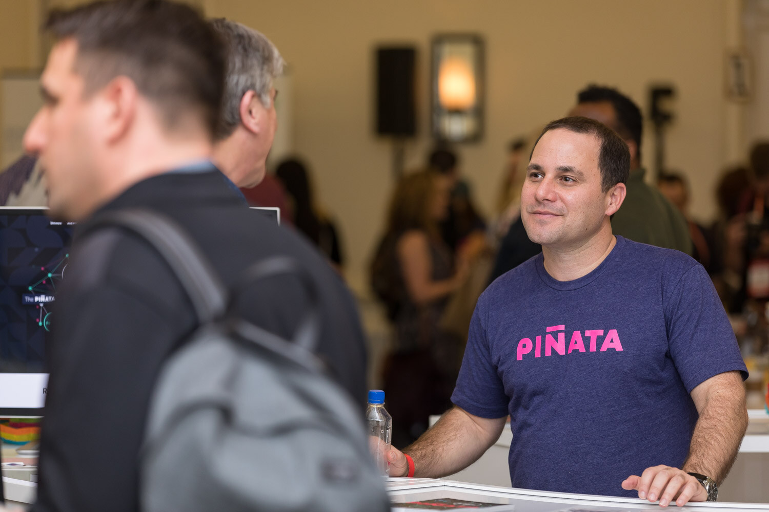 A man in a Piñata shirt smiles at his booth while an attendee in a backpack stands in front.
