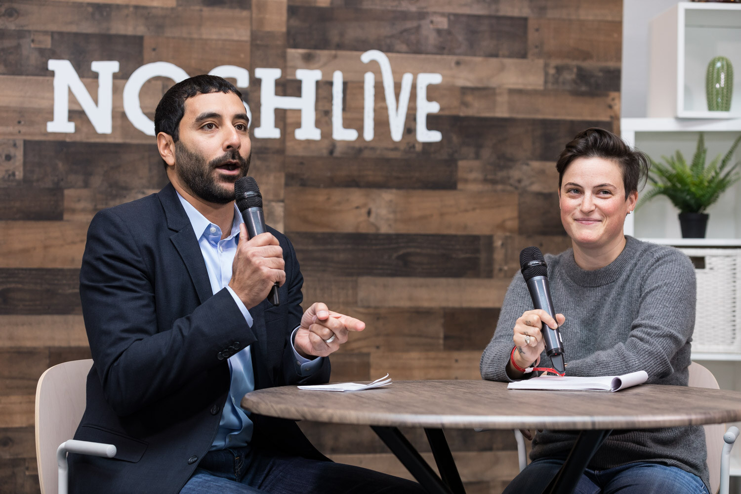 A host and a guest in a gray sweater talk at a round table in front of the Nosh Live wooden backdrop.