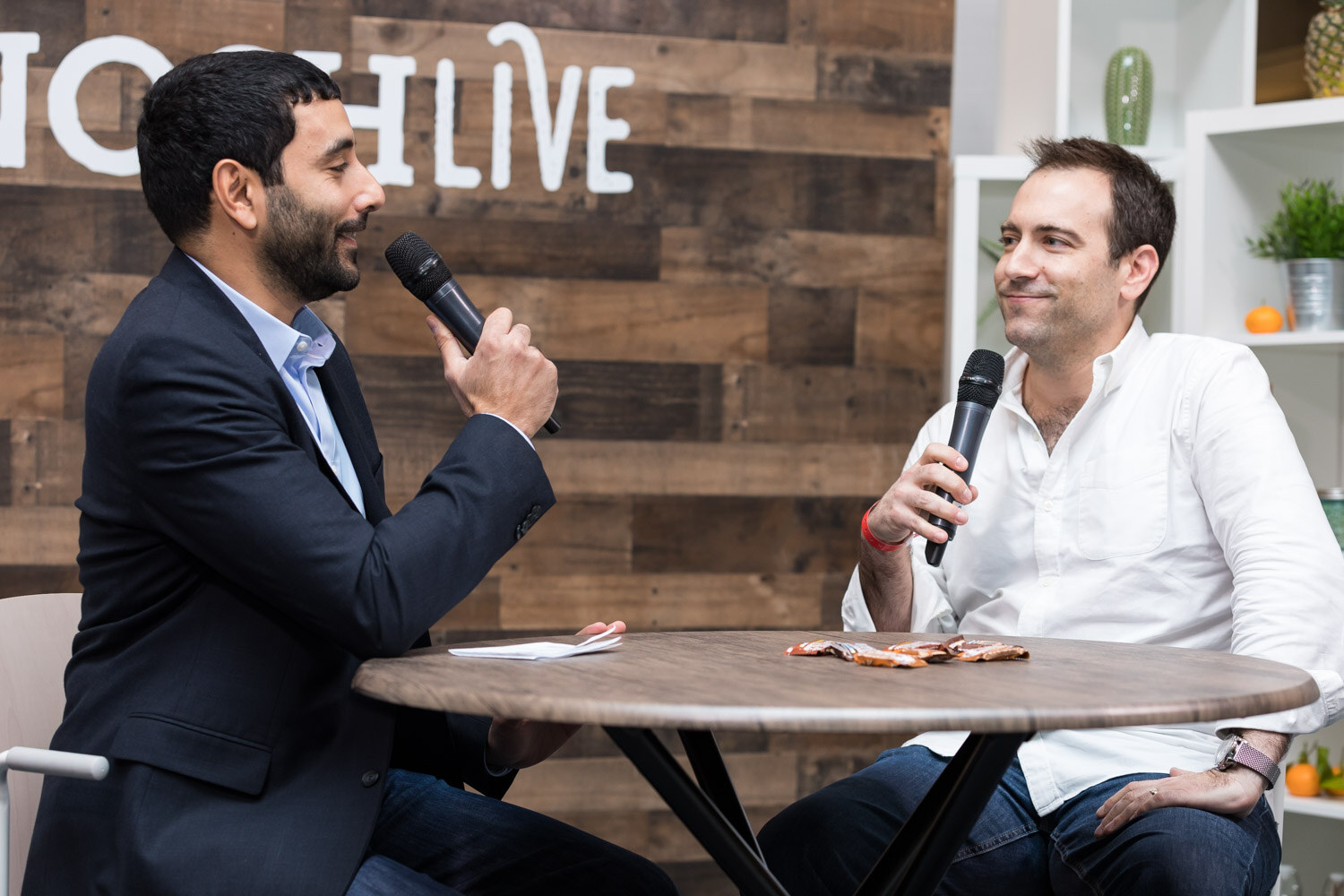 A host interviews a man in a white shirt at a round table in the livestream studio.