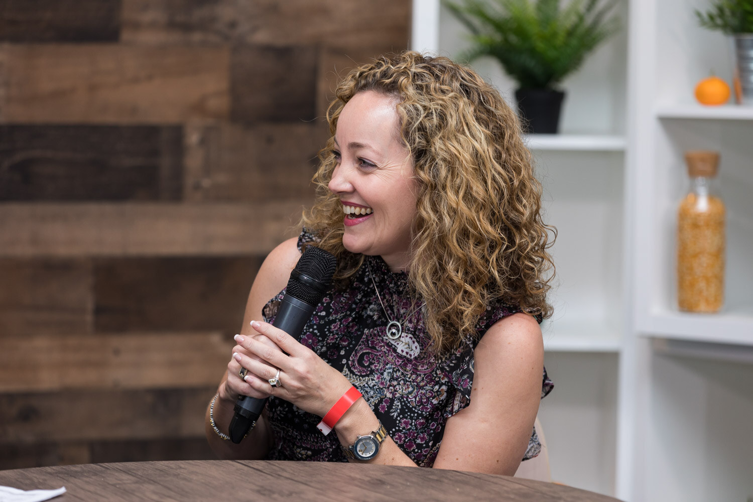 A woman with curly hair laughs while holding a microphone during a livestream interview.