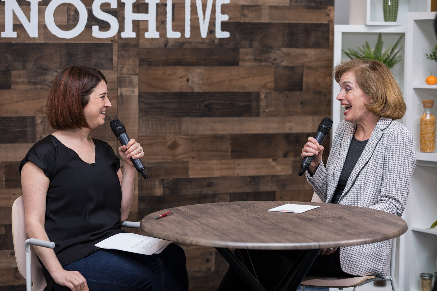 Two women talk at a round table in front of the Nosh Live sign in the livestream studio.