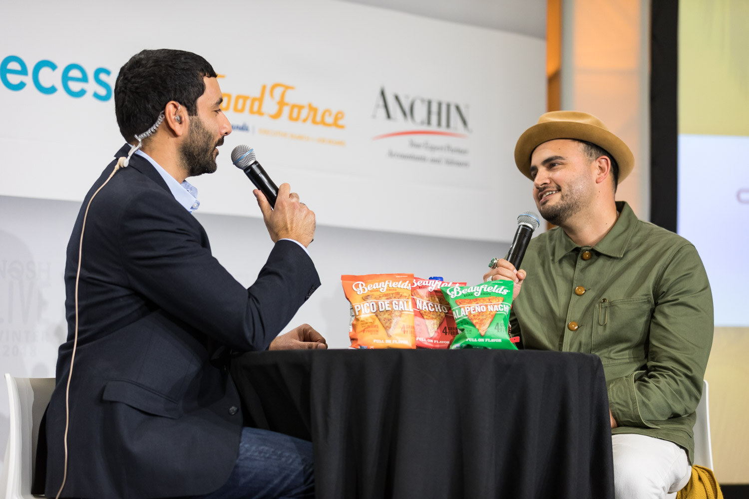A host interviews a man in an olive green shirt and tan hat on a stage with Beanfields chip bags on the table.