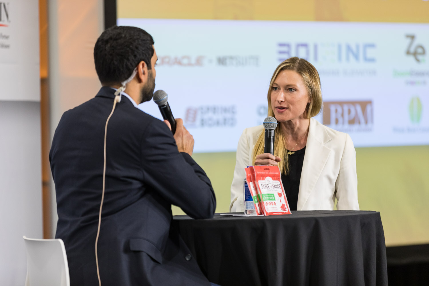 A host interviews a woman in a white blazer at a black-draped table on the livestream stage.