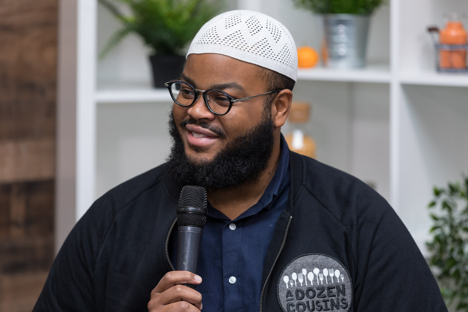 A smiling man in a white kufi and black jacket with an A Dozen Cousins patch holds a microphone.