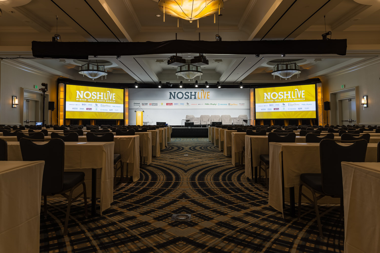 The main ballroom set for the conference with rows of tables facing the stage and Nosh Live screens.
