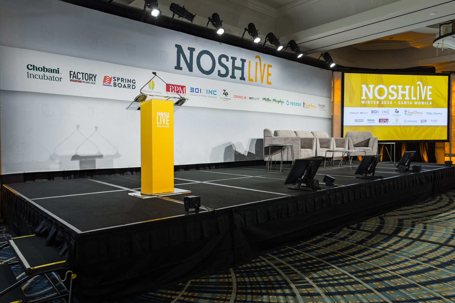 The main stage with a yellow lectern and gray armchairs under the Nosh Live banner and sponsor logos.