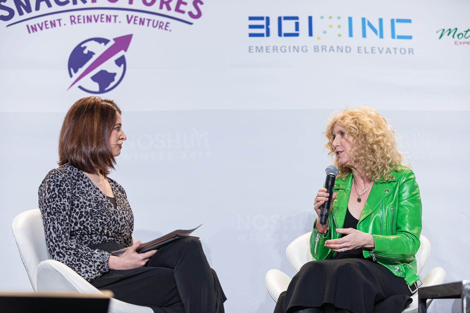 A woman in a green jacket speaking into a microphone to a seated interviewer holding papers