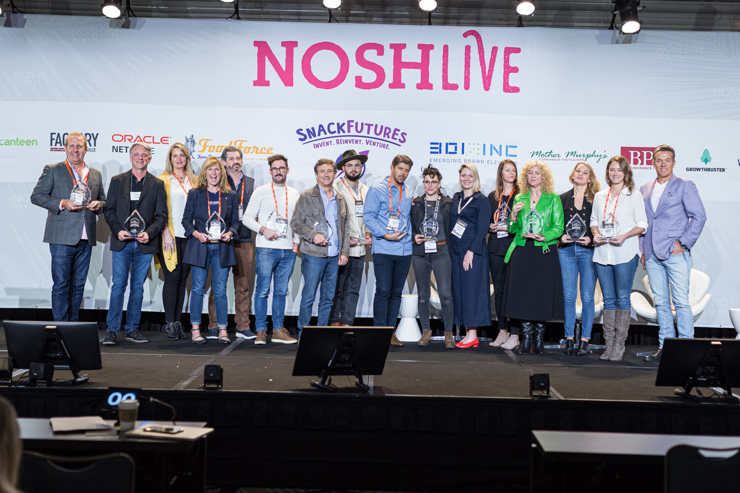 A large group of people standing in a row on a stage holding glass award trophies in front of a Nosh Live backdrop