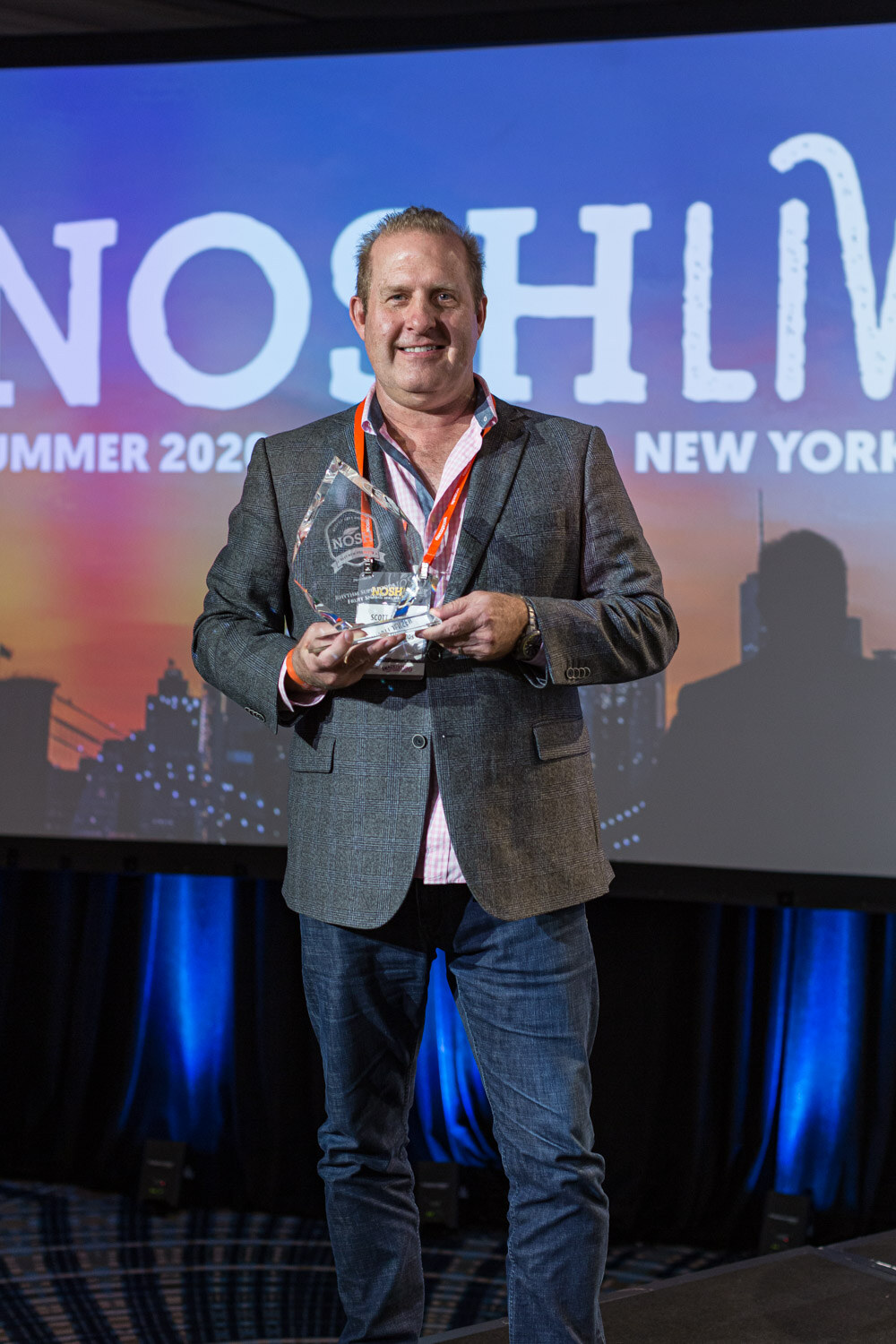 A man in a gray blazer holds a glass trophy in front of a Nosh Live banner