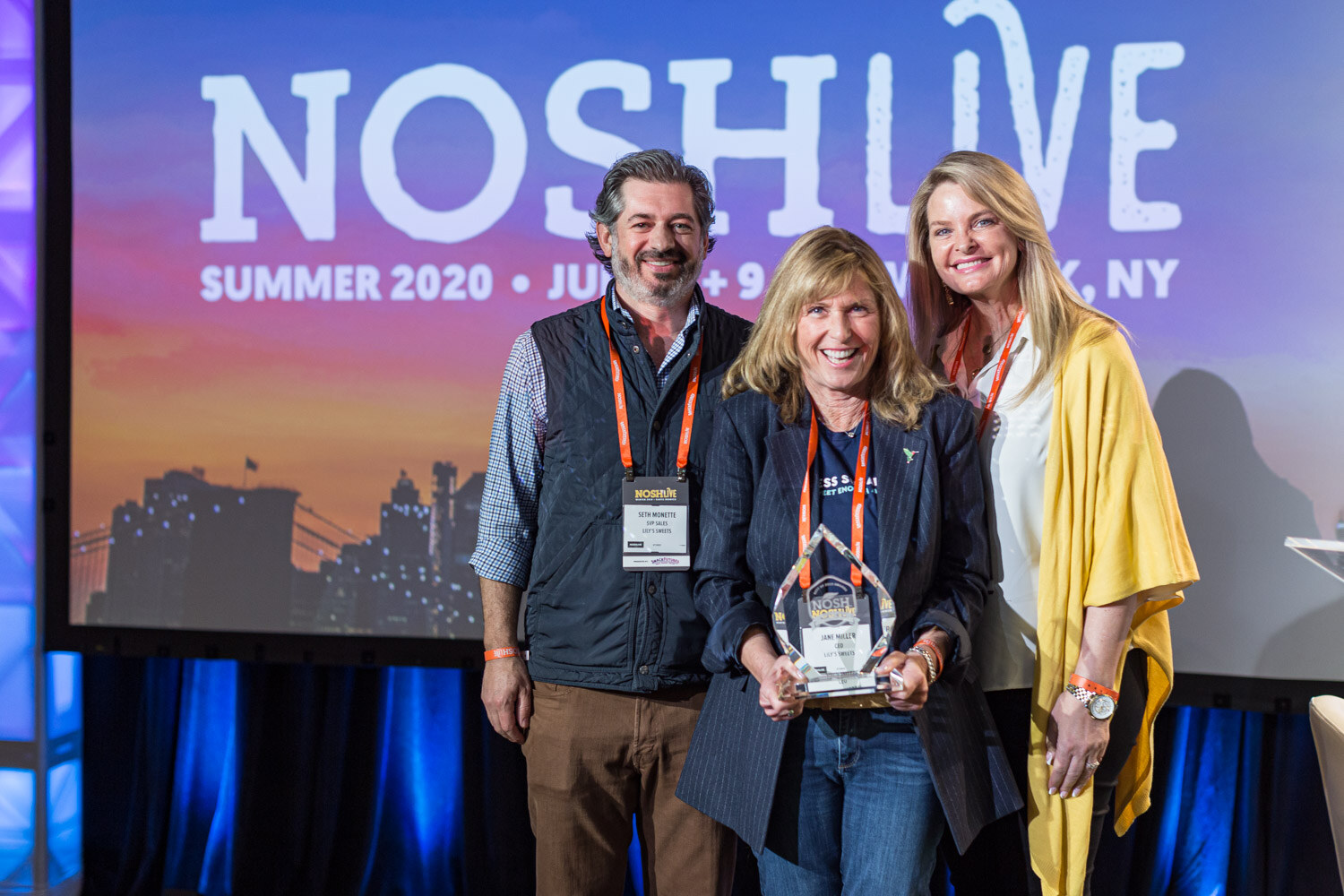 Two women and a man pose together, one woman holding a glass trophy, in front of a Nosh Live backdrop