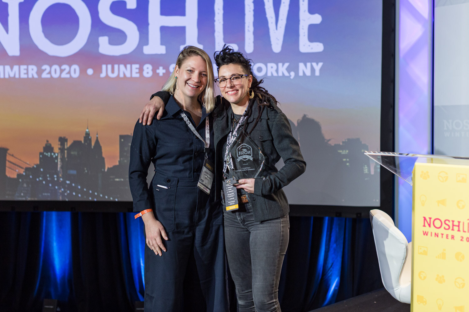 Two women in dark clothes hug on stage and hold glass trophies beside a yellow Nosh Live lectern