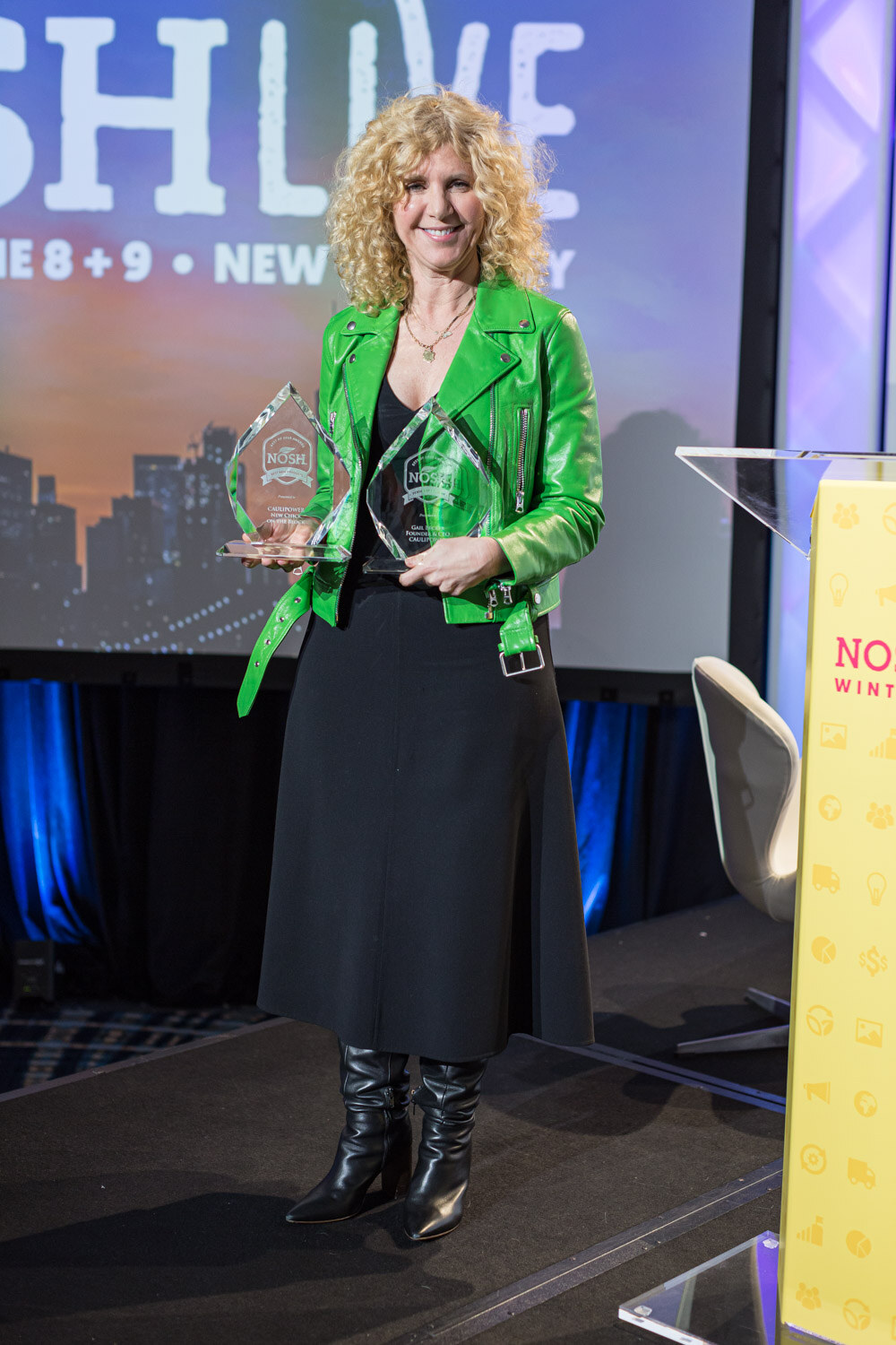 A woman in a bright green jacket holds two glass awards on the Nosh Live stage