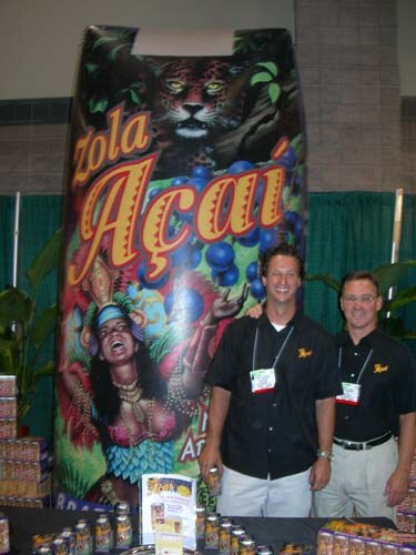 Two men in black shirts and badges stand in front of a tall Zola Açaí display showing a jaguar and a woman in costume, with product bottles and boxes on the table.