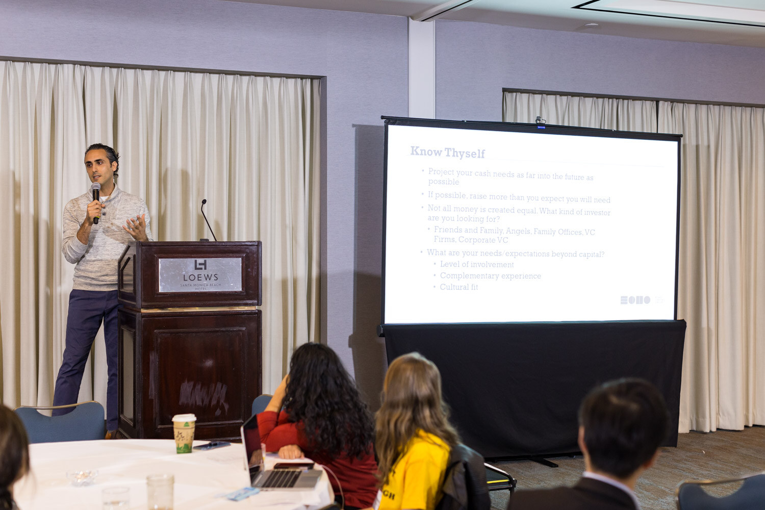A man speaking into a microphone at a hotel lectern beside a projector screen showing a slide