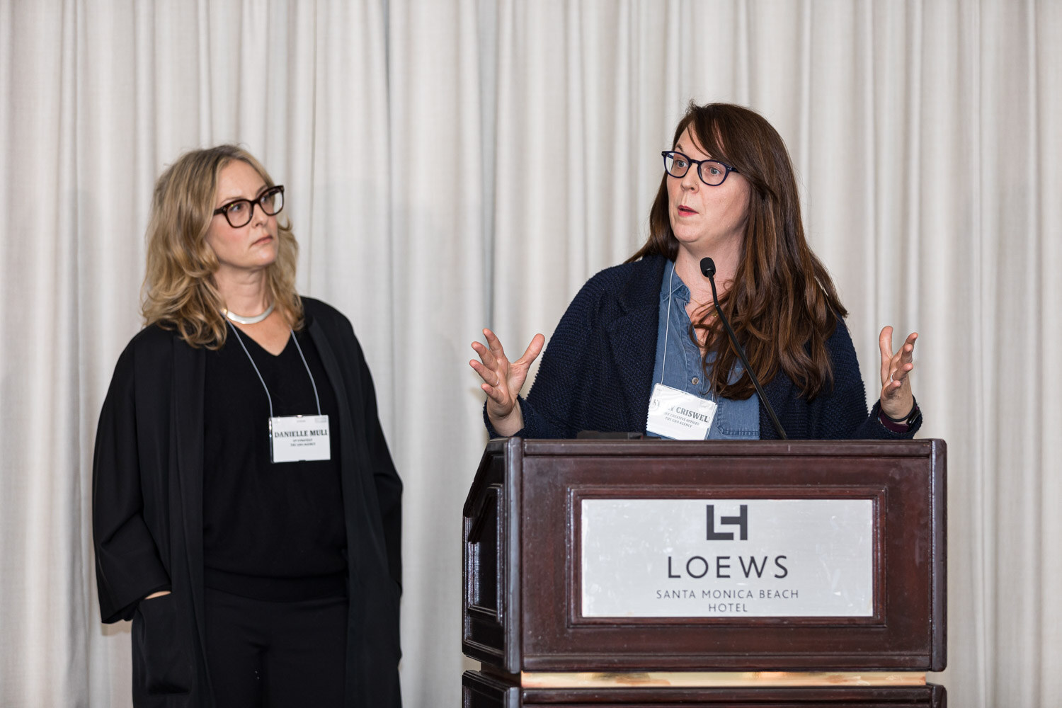 Two women stand at a Loews lectern, one speaking with hands raised, in front of cream curtains