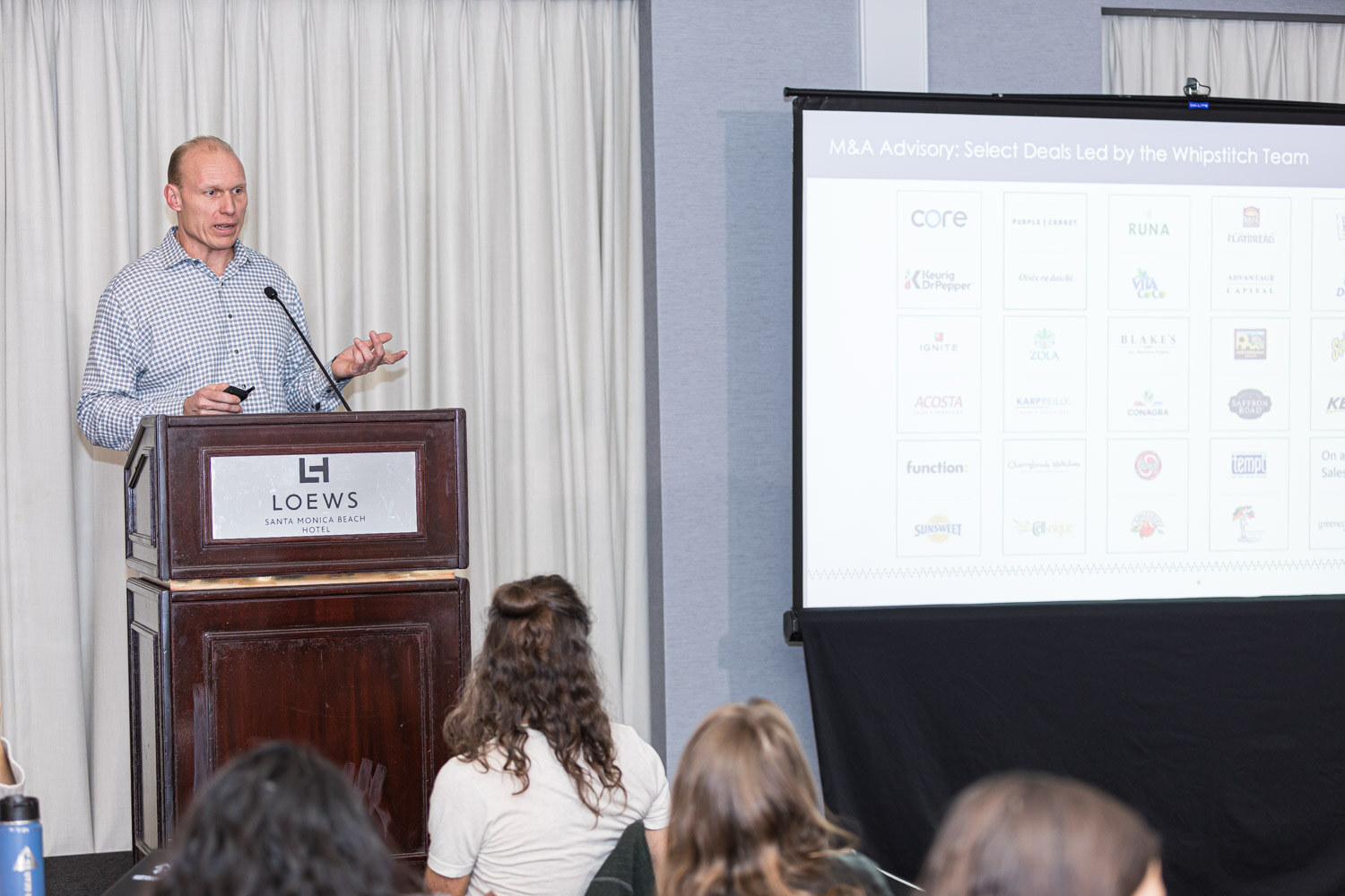 A man at a lectern beside a screen showing M and A advisory deals with brand logos