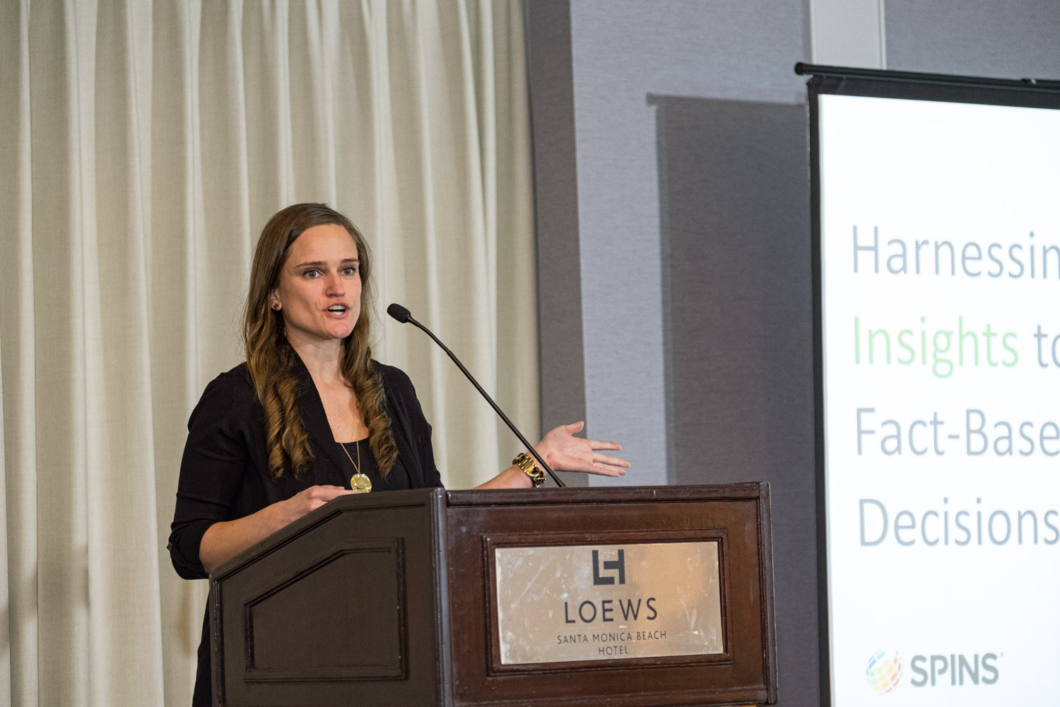 A woman speaking at a Loews lectern beside a SPINS banner reading Harnessing Insights to Fact-Based Decisions