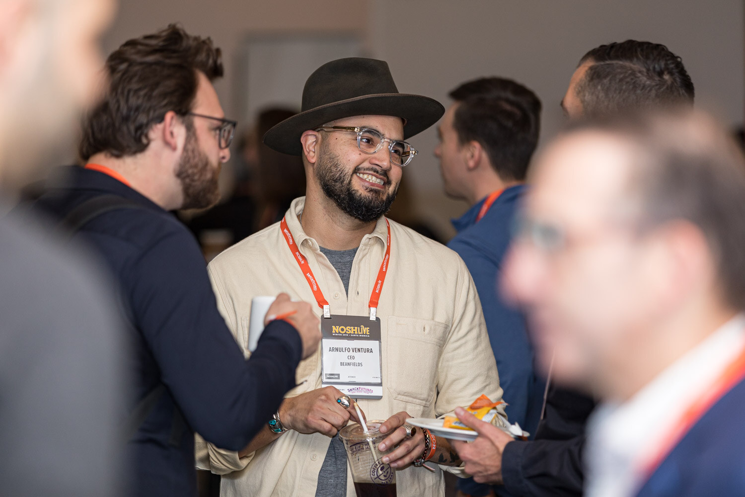 A smiling man in a hat and glasses with an orange lanyard talking with other attendees