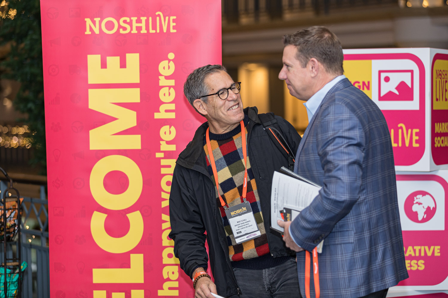 Two men with lanyards in conversation next to a pink and yellow Nosh Live welcome banner