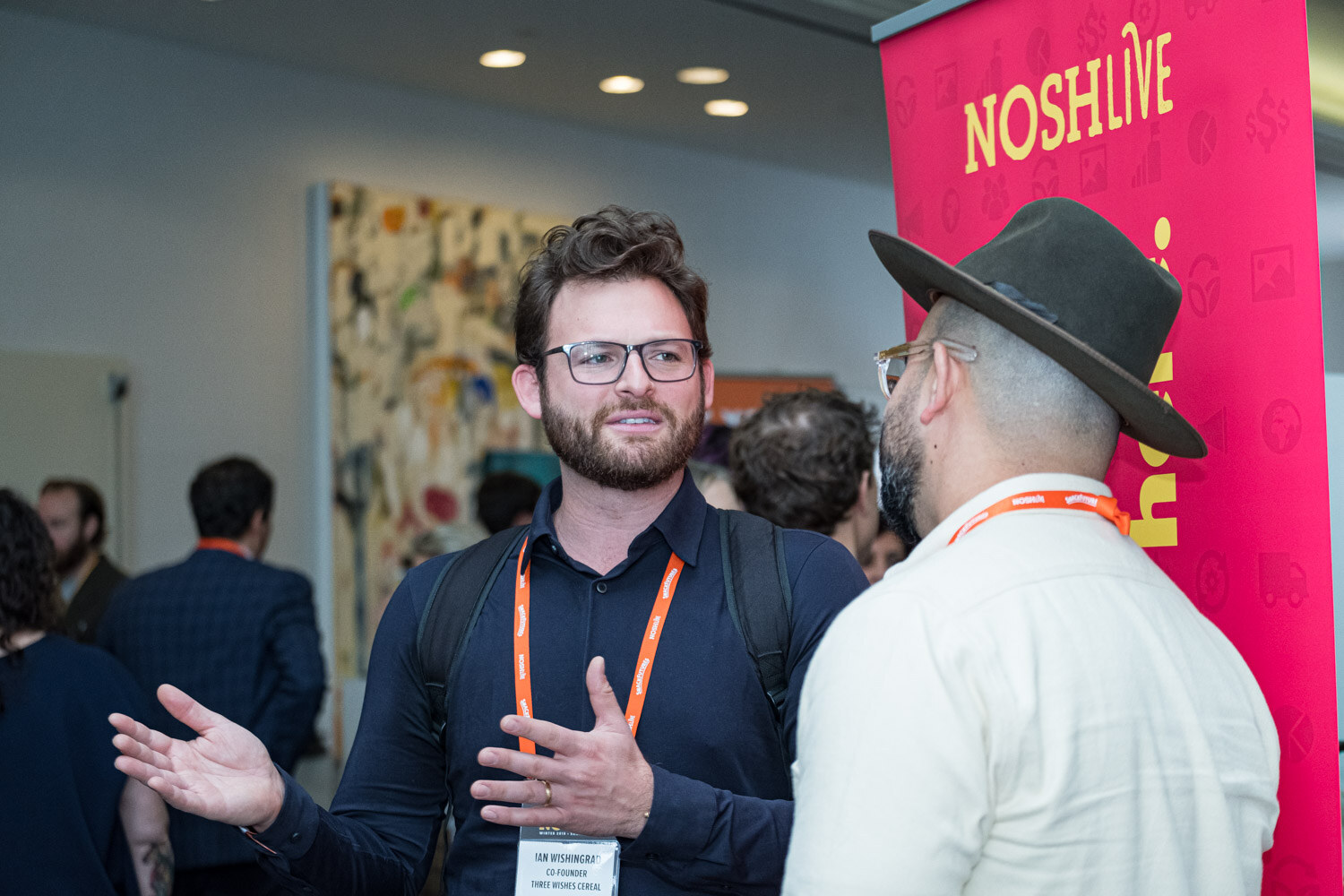A bearded man in glasses gestures while talking to a man in a hat beside a pink Nosh Live banner