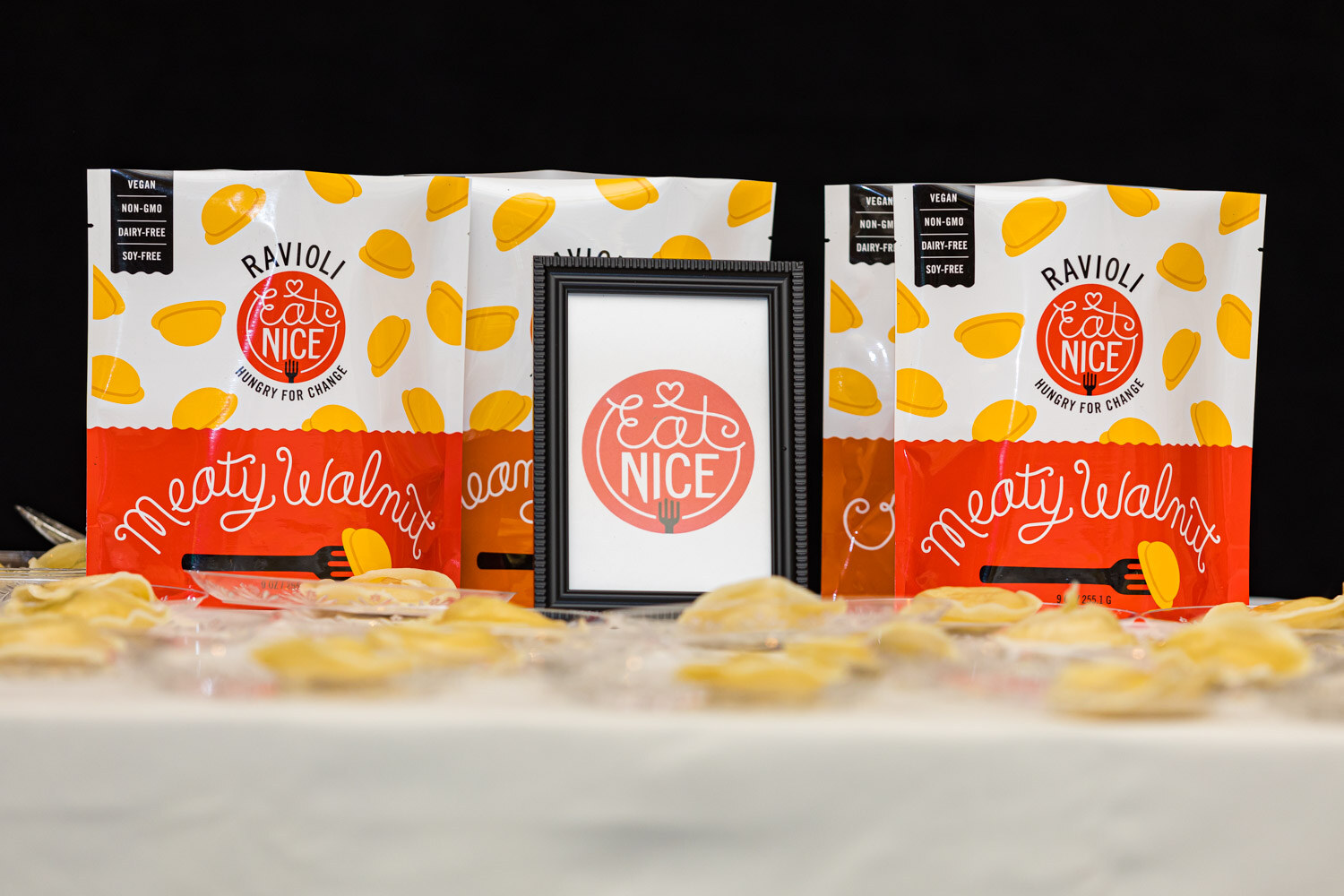 Packages of Eat Nice Meaty Walnut Ravioli beside a framed Eat Nice sign