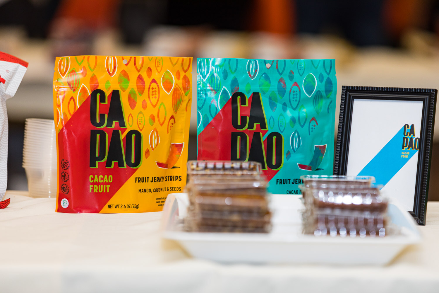 Bags of Ca Pao Fruit Jerkystrips in yellow and teal beside a tray of samples
