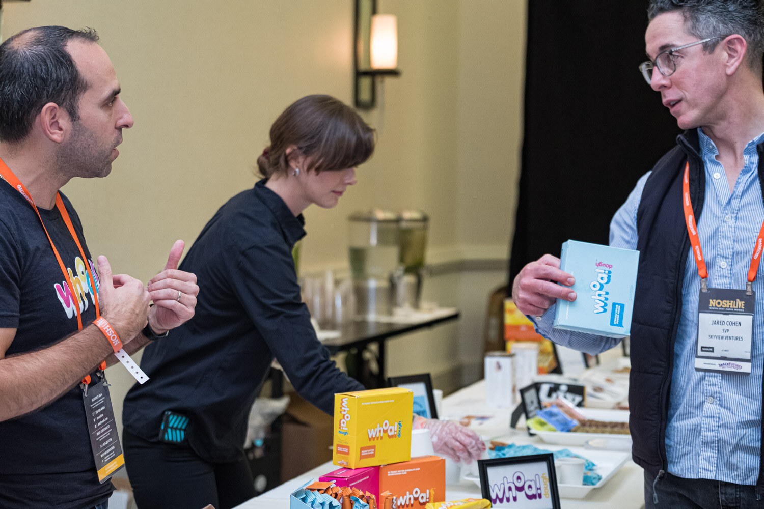 A man in a black Whoa Dough shirt and a woman setting out product while an attendee holds a box