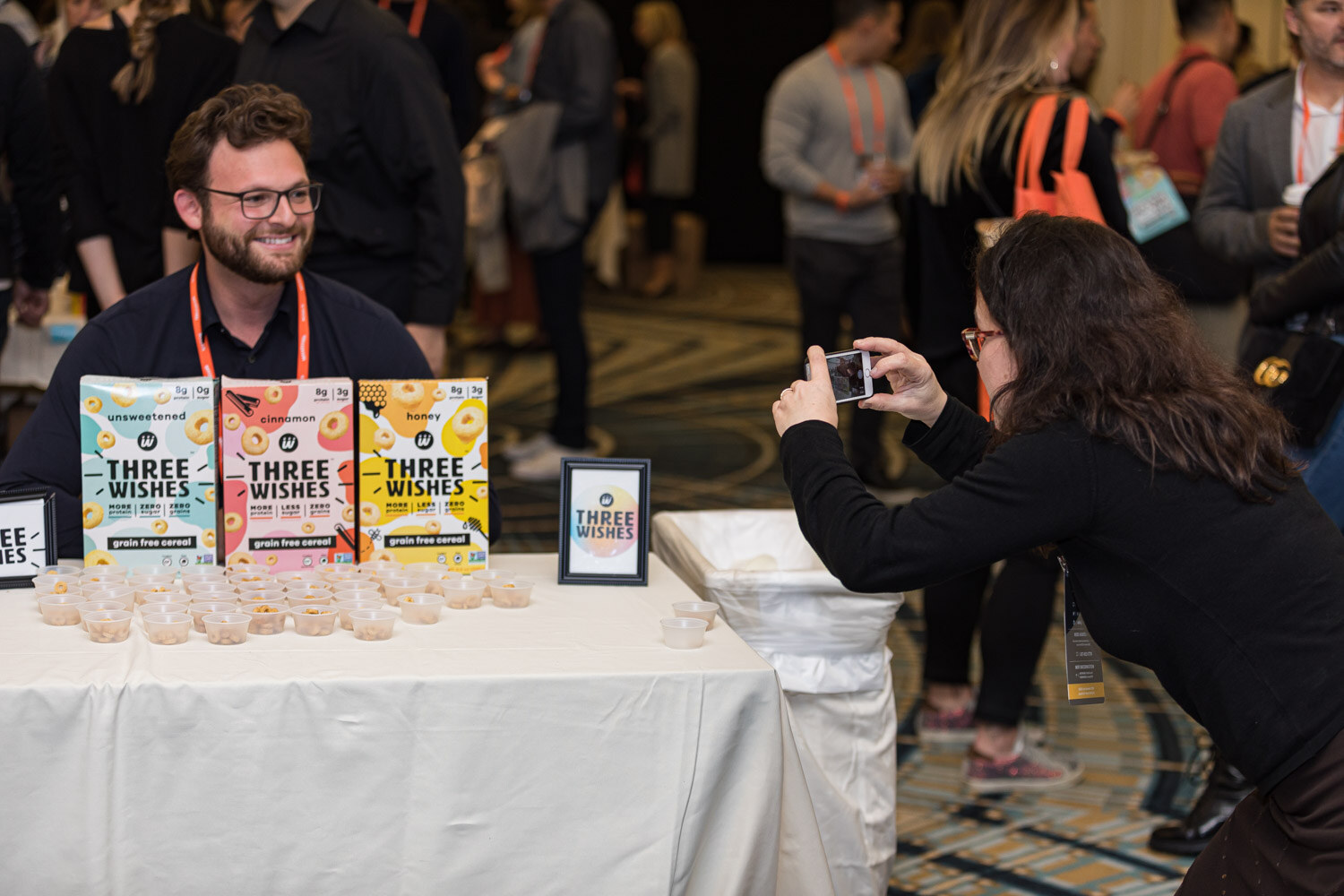 A smiling man with glasses sits behind Three Wishes cereal boxes while an attendee photographs the table