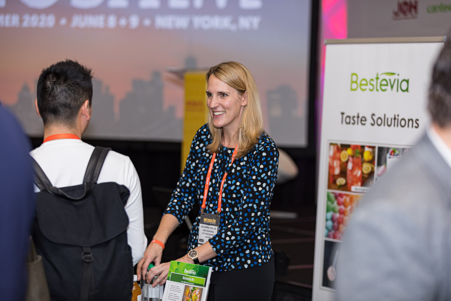 A smiling woman in a polka-dot top beside a Bestevia Taste Solutions banner