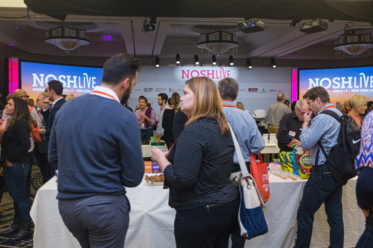 Attendees crowd around sampling tables in the ballroom with Nosh Live screens behind