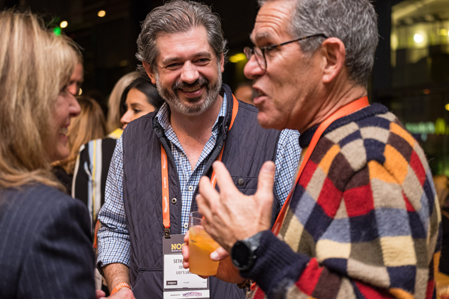 A smiling bearded man in a vest talking with a man in a checked sweater at a restaurant party, both wearing orange lanyards